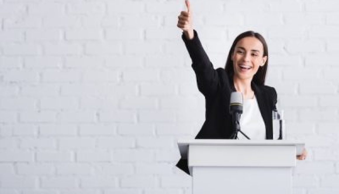 happy, attractive lecturer showing thumb up while standing on podium tribune