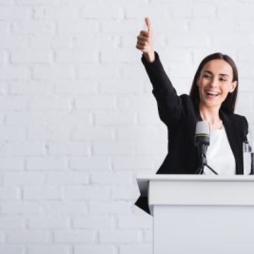 happy, attractive lecturer showing thumb up while standing on podium tribune